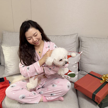 Woman in pink pajamas holding a small white dog on a couch with a gift box and decorative pillow.